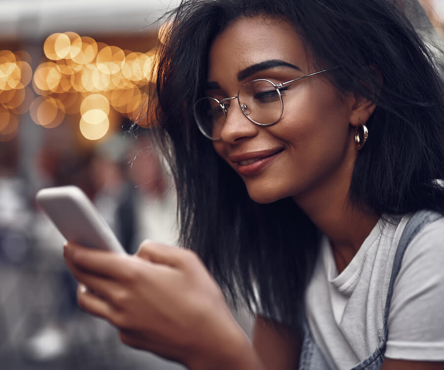 Jeune femme souriante utilisant une application mobile qui lui permet d'être connecté à distance et de communiquer. Fond de lumière urbain calme.