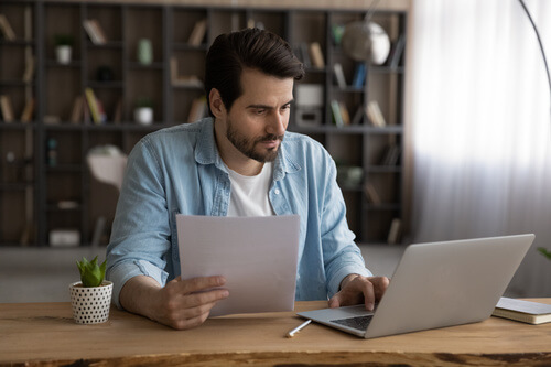 Un homme tient une lettre dans la main droite et consulte son ordinateur de bureau avec la main gauche.