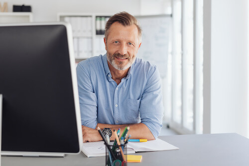 un homme dans un bureau d'entreprise se tient sur ses coudes devant son ordinateur et sourit.